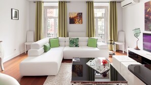 A modern living room with a white sectional sofa, green cushions, a glass coffee table, and a large window with green curtains. - Lisbon Rentals Chiado (Lisbon)