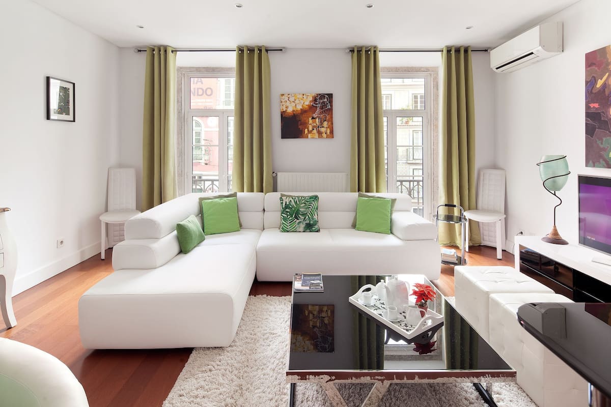 A modern living room with a white sectional sofa, green cushions, a glass coffee table, and a large window with green curtains.