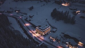 Paesaggio innevato con un grande edificio dotato di numerose finestre e un tetto piatto, circondato da alberi coperti di neve e alcuni edifici più piccoli.
