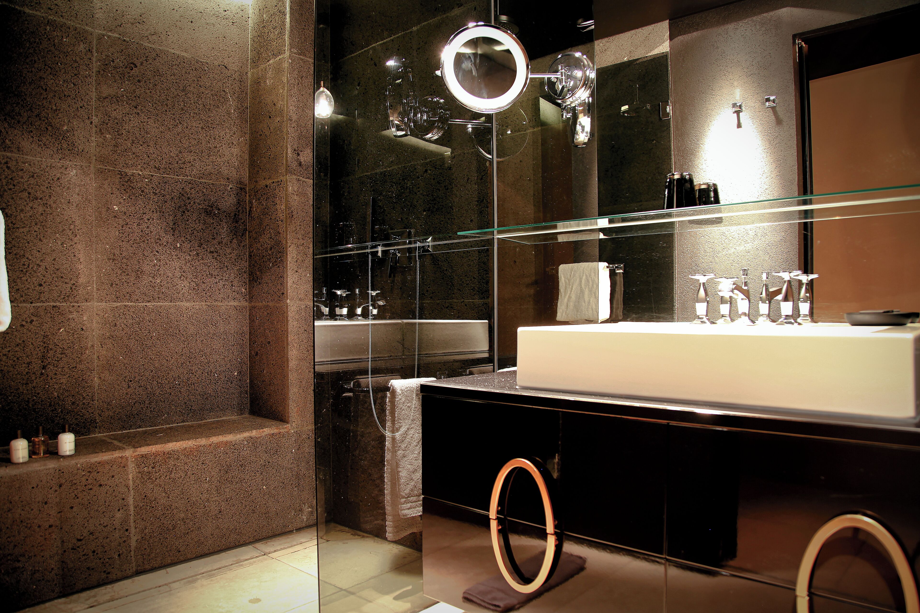 A modern bathroom with a glass-enclosed shower, a large mirror with a ring light, and a vanity with a white sink and multiple bottles.