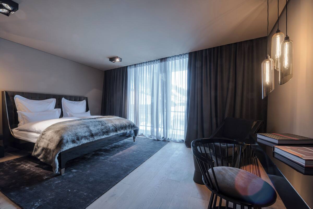 A modern bedroom with a large bed, a desk, and a chair. There is a window with curtains and a ceiling light fixture.