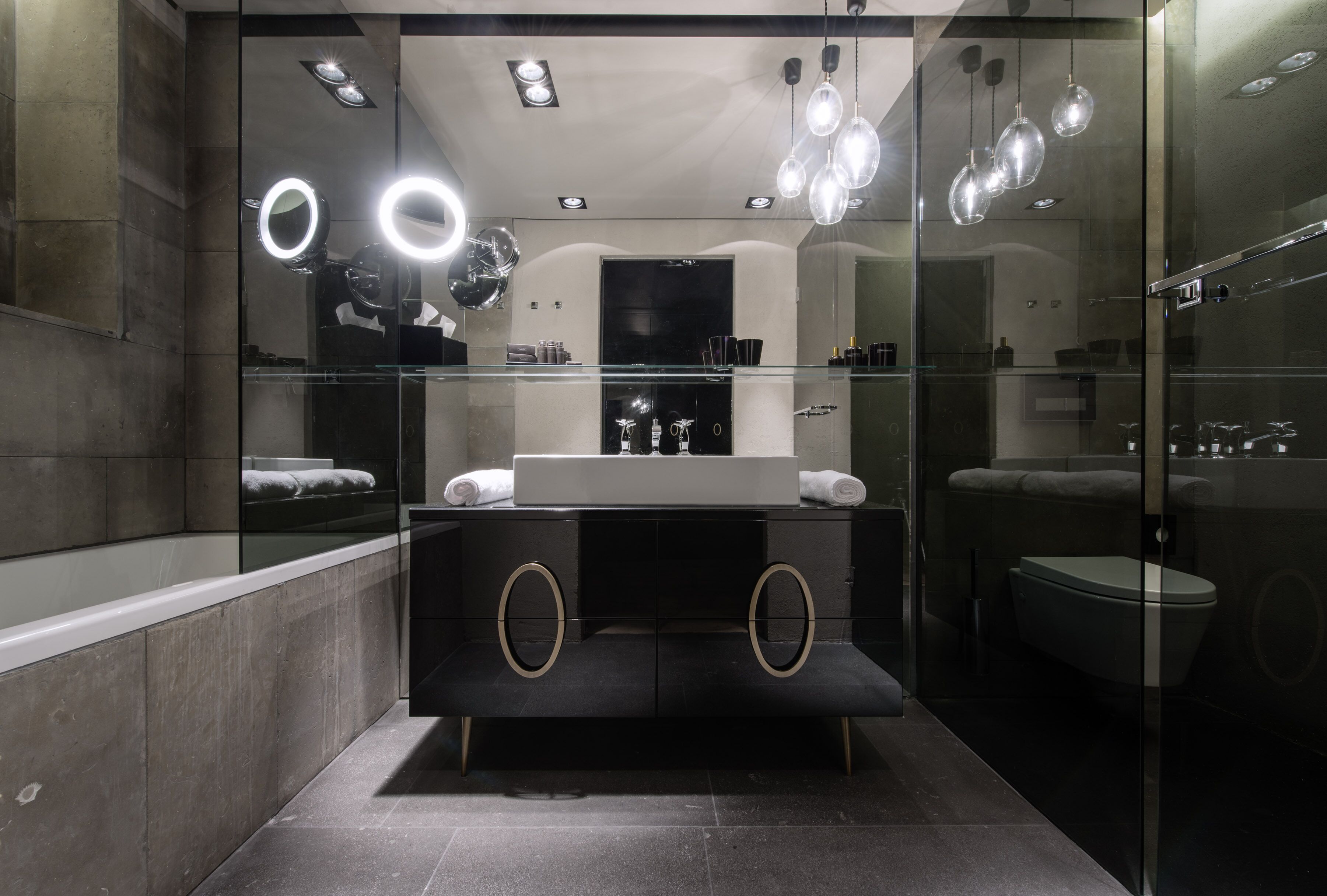 A modern bathroom with a glass-enclosed vanity, a large mirror with circular lights, and a glass shower enclosure.