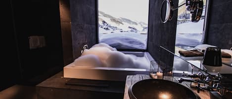 Une salle de bain moderne dotée d’une grande baignoire, d’un meuble-lavabo en bois et offrant une vue sur un paysage montagneux enneigé par le biais d’une grande fenêtre.