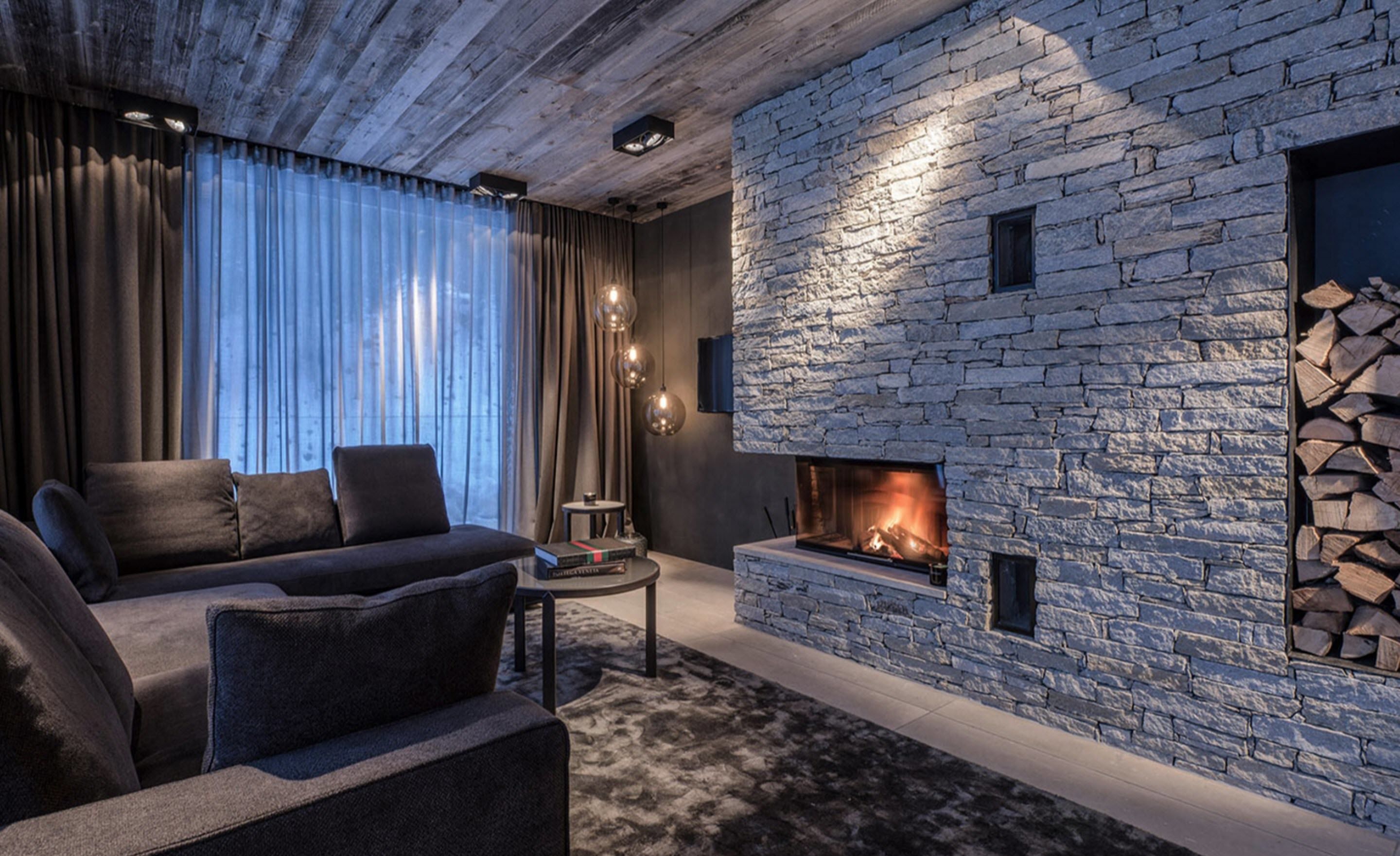 A modern living room with a stone fireplace, dark furniture, and a view of the outdoors through large windows.