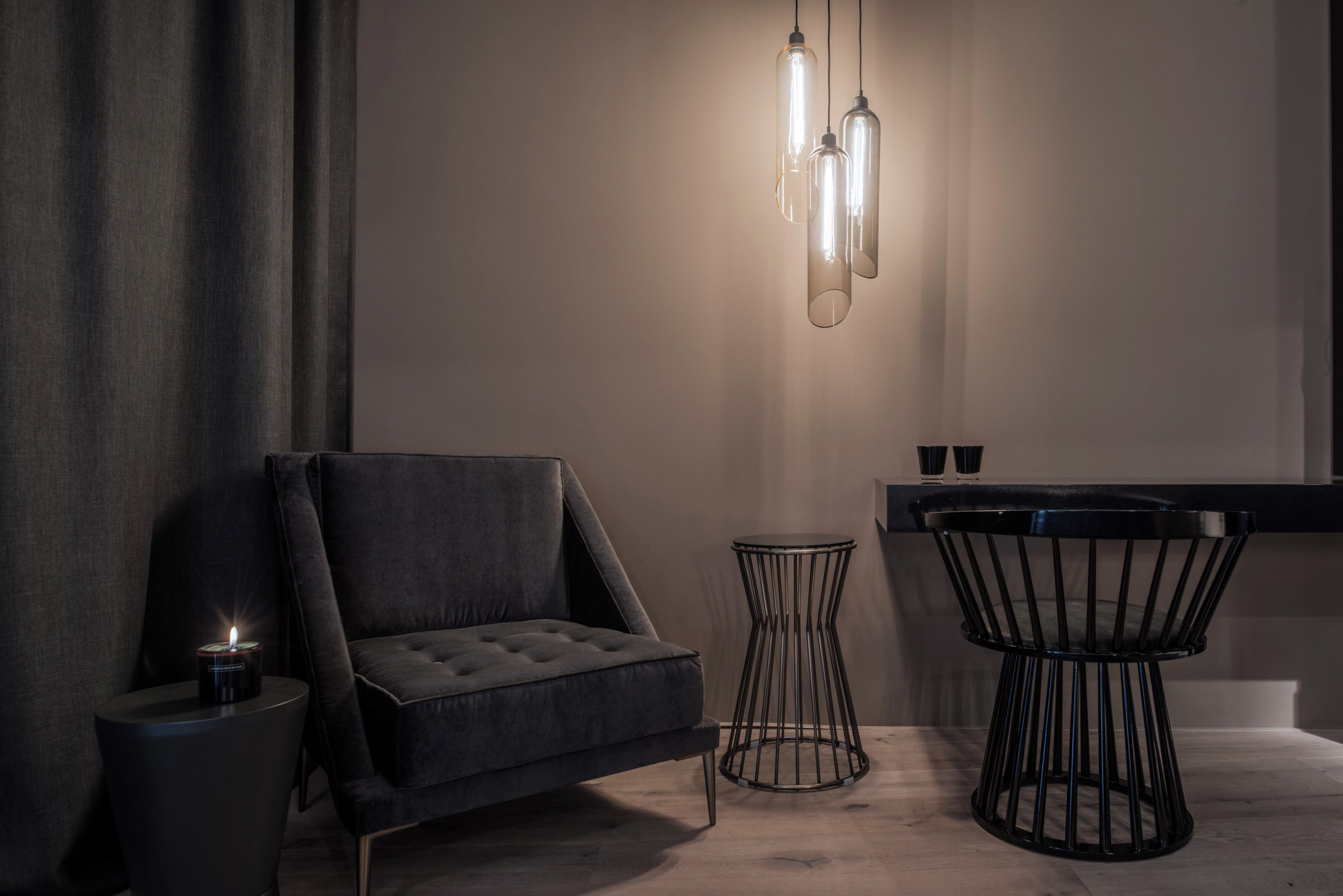 A modern room with a dark grey sofa, a round black side table with a candle, a black metal chair, and a hanging light fixture.