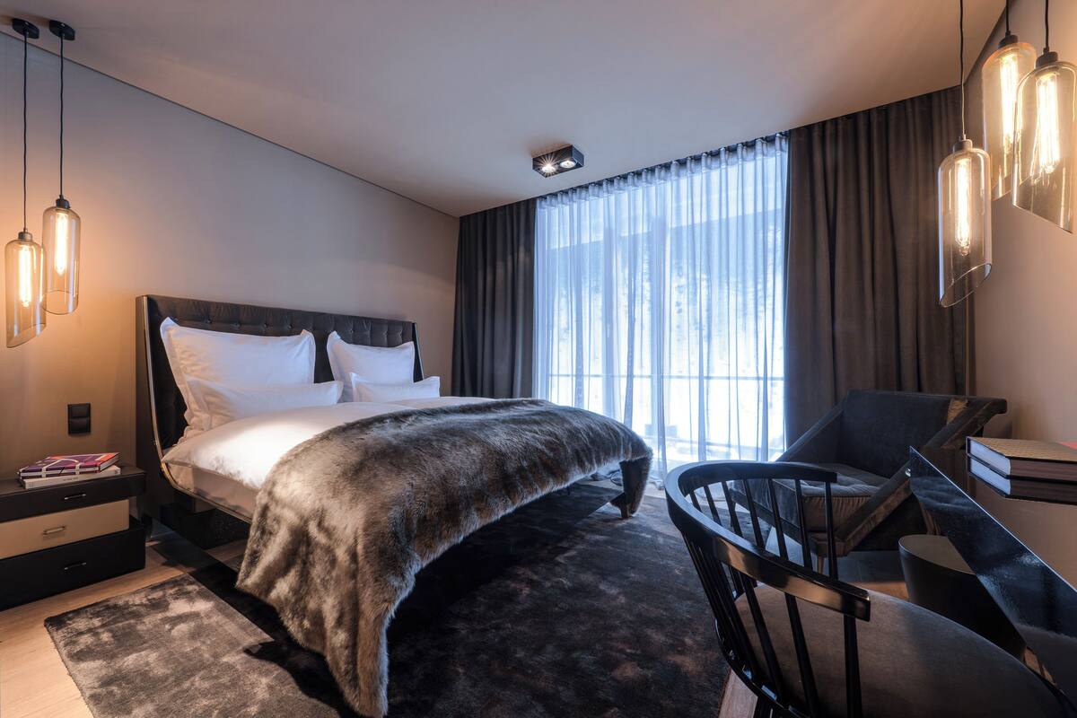 A modern bedroom with a large bed, a desk, and a chair. There are hanging lights and a window with curtains.