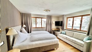 A modern hotel room with a large bed, a sofa, a TV, and large windows. - Haus Schrofenstein (Lech am Arlberg)