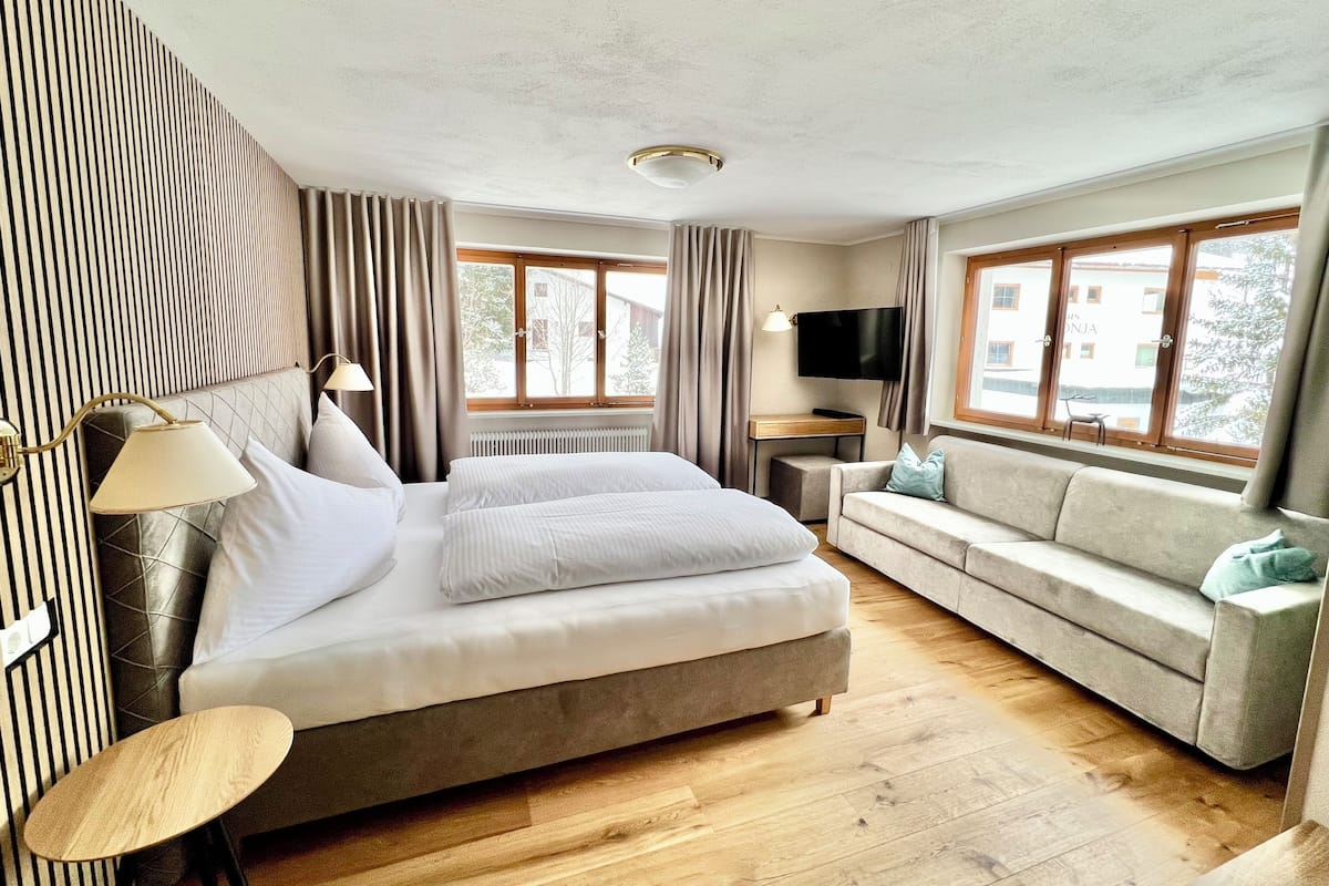 Una habitación de hotel moderna con una cama grande, un sofá, un televisor y ventanas amplias.