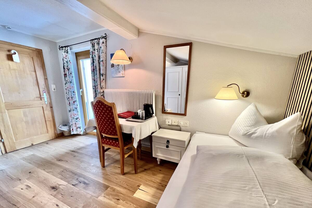 Habitación de hotel con piso de madera, un escritorio con silla, cama con ropa blanca, mesita de noche, espejo y ventana con cortinas.