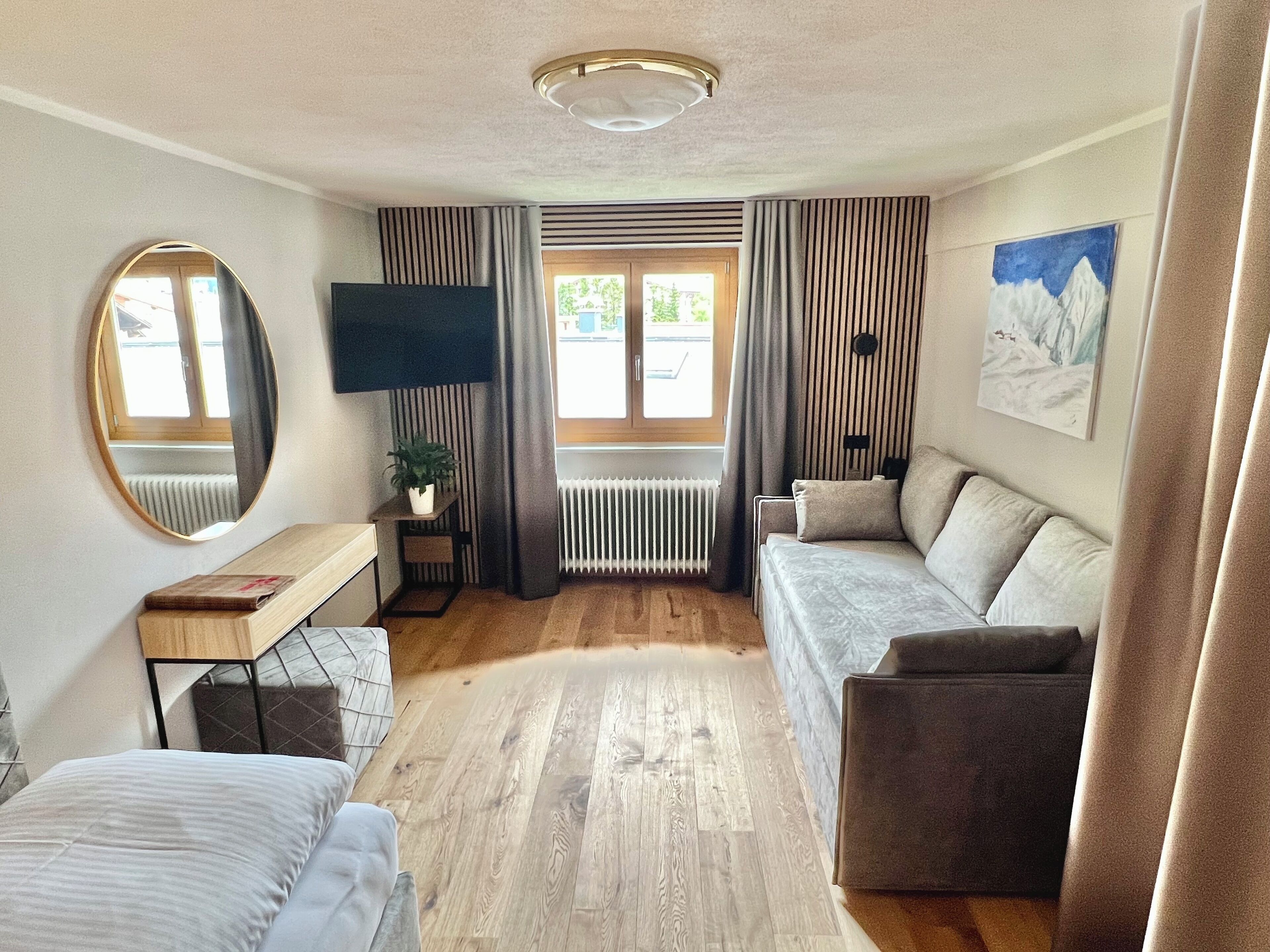 Una habitación de hotel moderna con suelo de madera, un sofá, una cama, un escritorio con planta, un televisor y un cuadro en la pared.