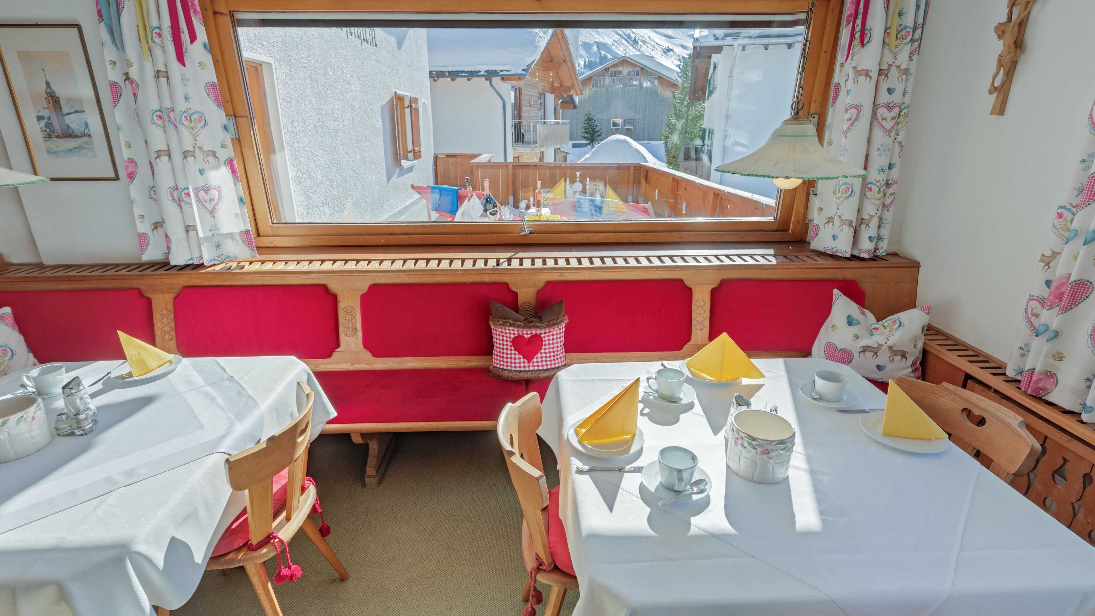 A cozy dining area with a wooden bench and tables, a view of a snowy mountain, and a window with curtains.
