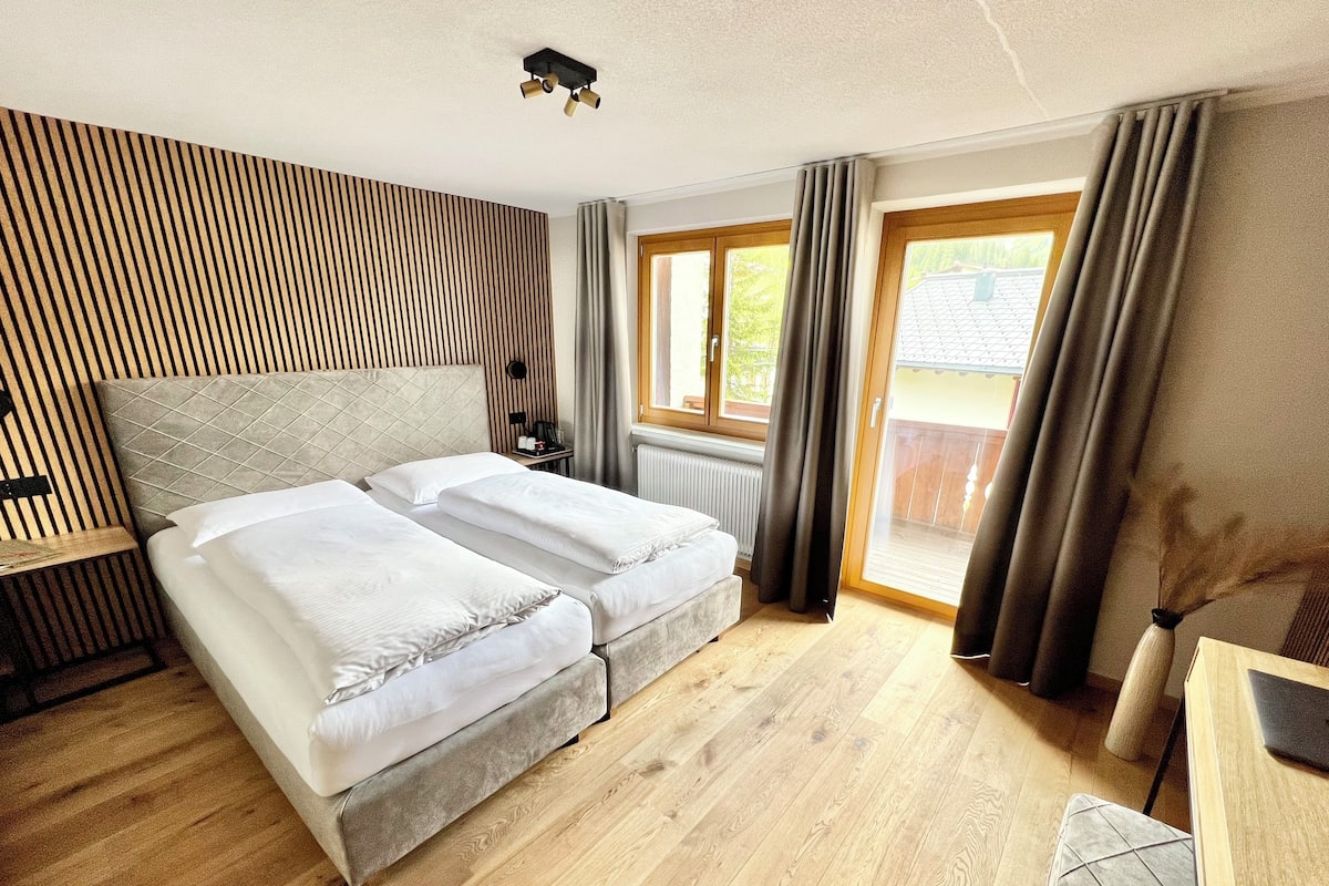 Habitación de hotel moderna con suelo de madera, una cama grande con ropa blanca, un escritorio pequeño y vistas al exterior a través de una puerta corrediza de vidrio.