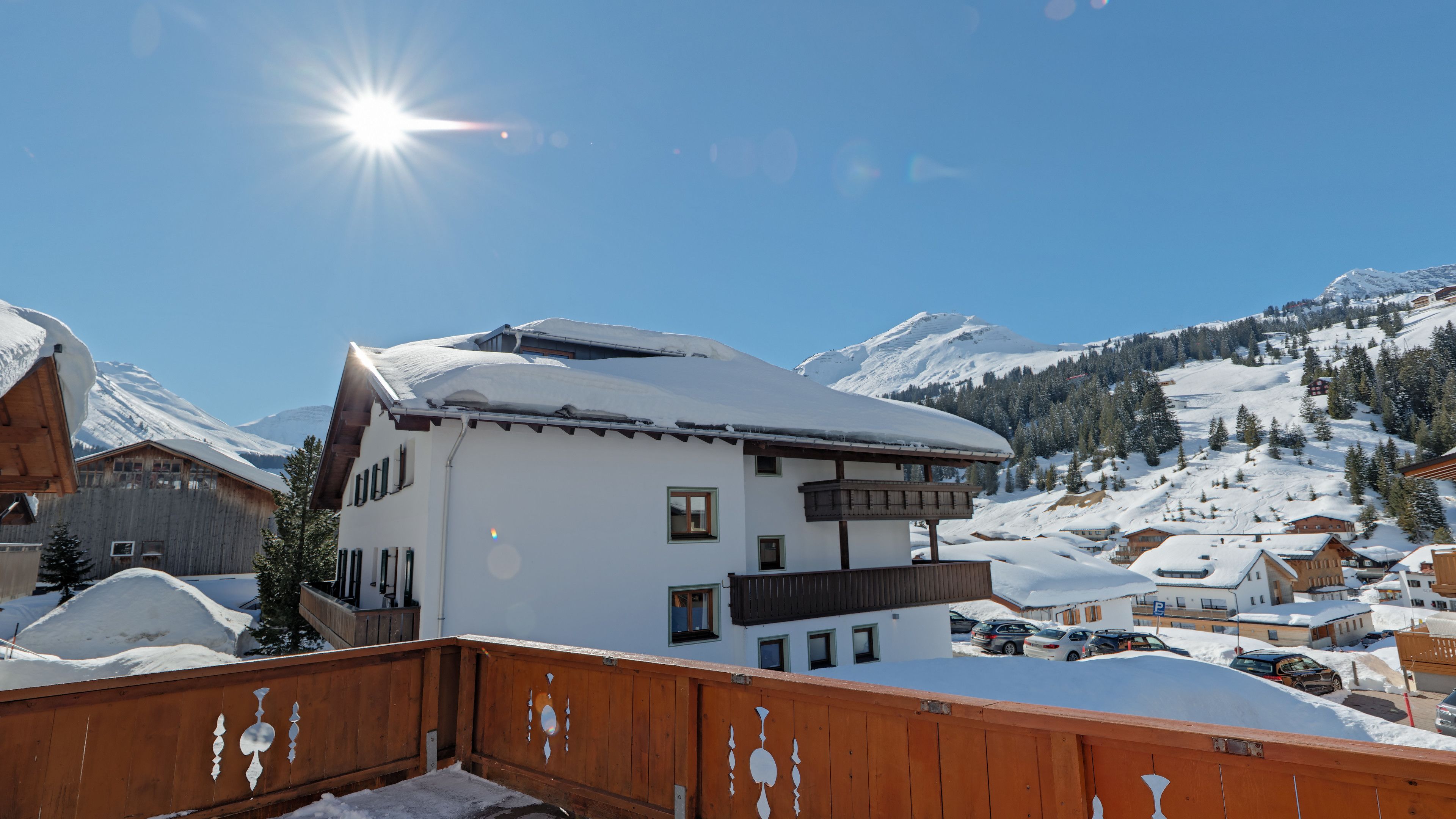 A snow-covered mountain village with a prominent white building featuring balconies and a wooden fence.