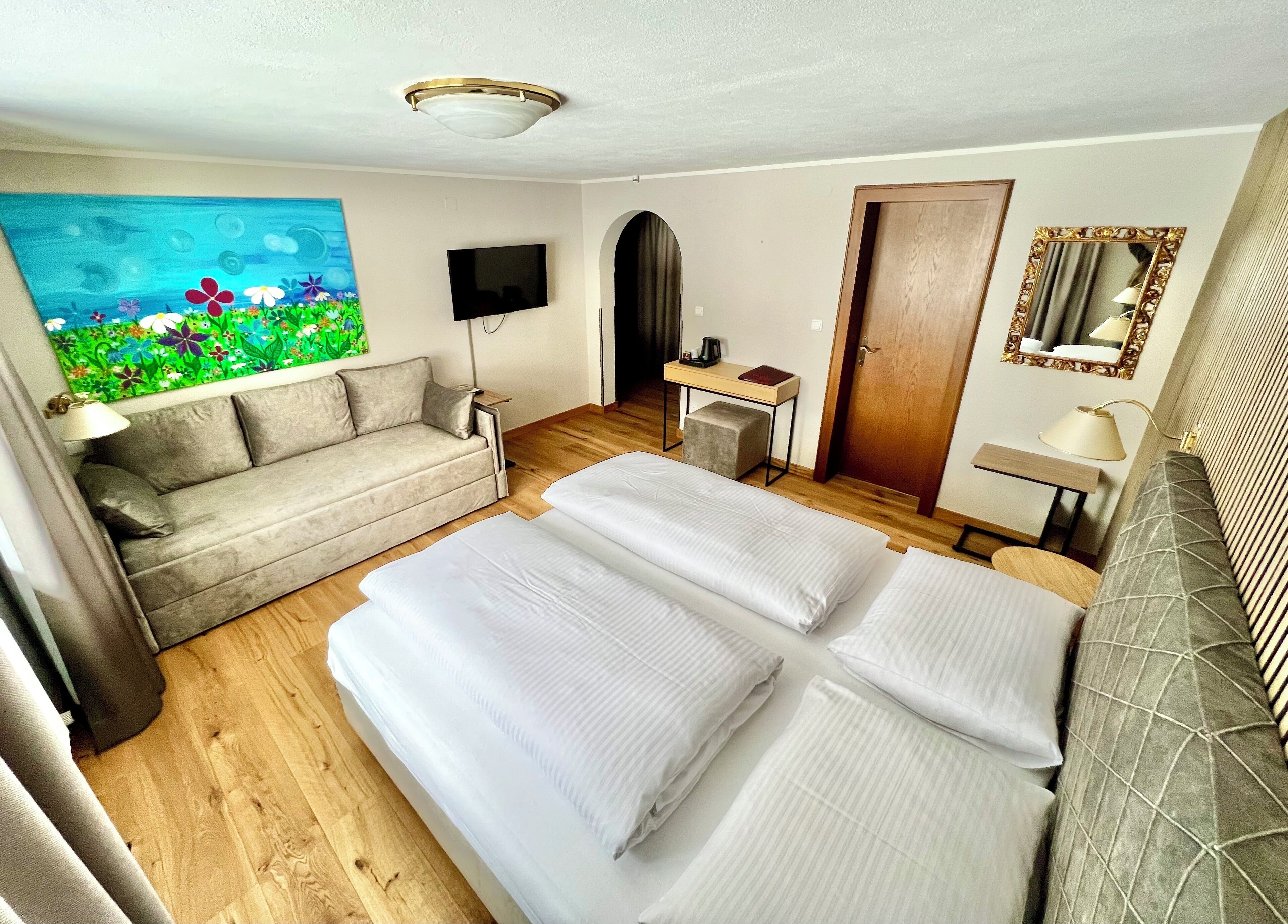 A hotel room with a bed, a sofa, a desk, a TV, a painting, and a door.