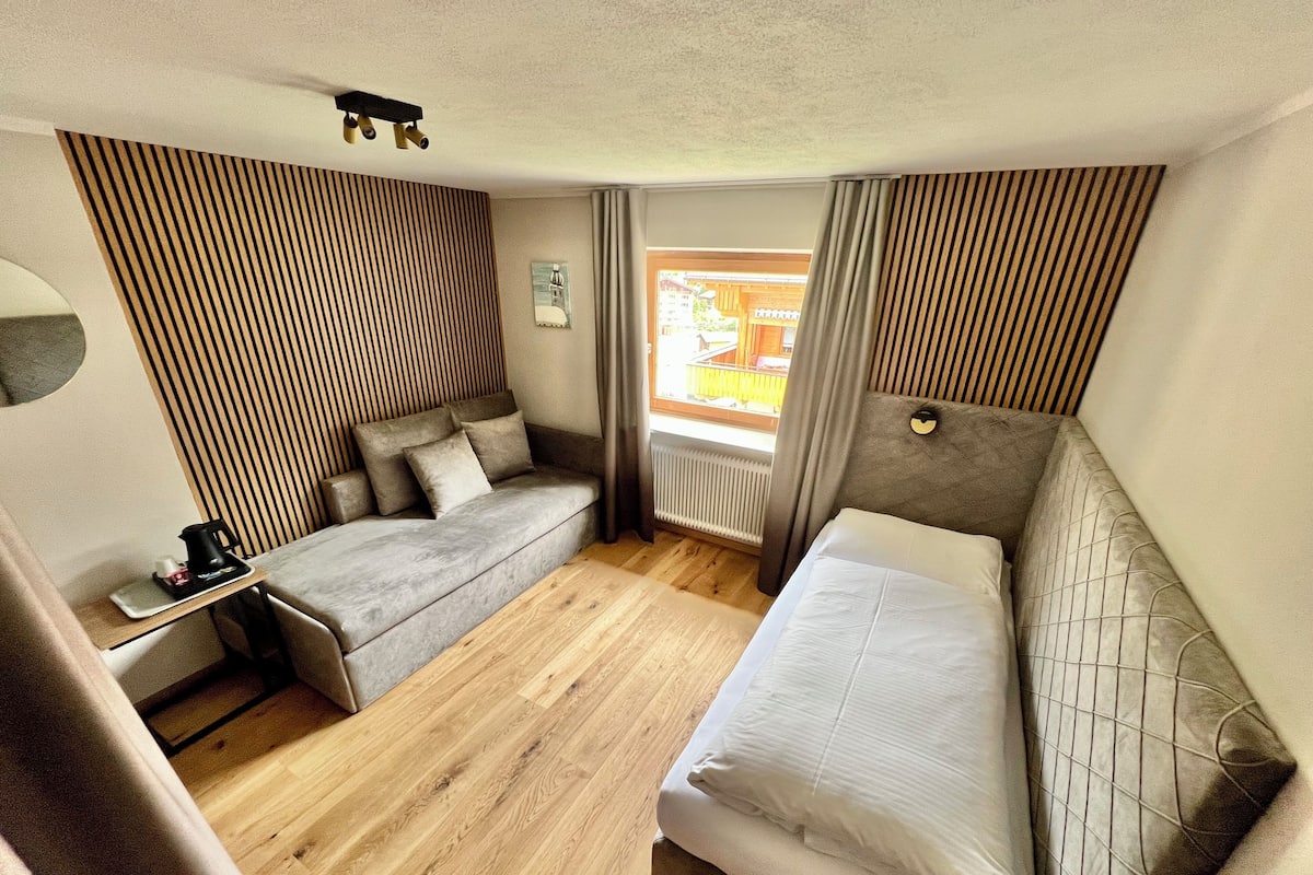 Habitación de hotel compacta con sofá, cama, una mesita con hervidor y ventana con cortinas.