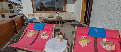 A terrace with sun loungers, a table with a bottle of wine, and a view of a mountain.