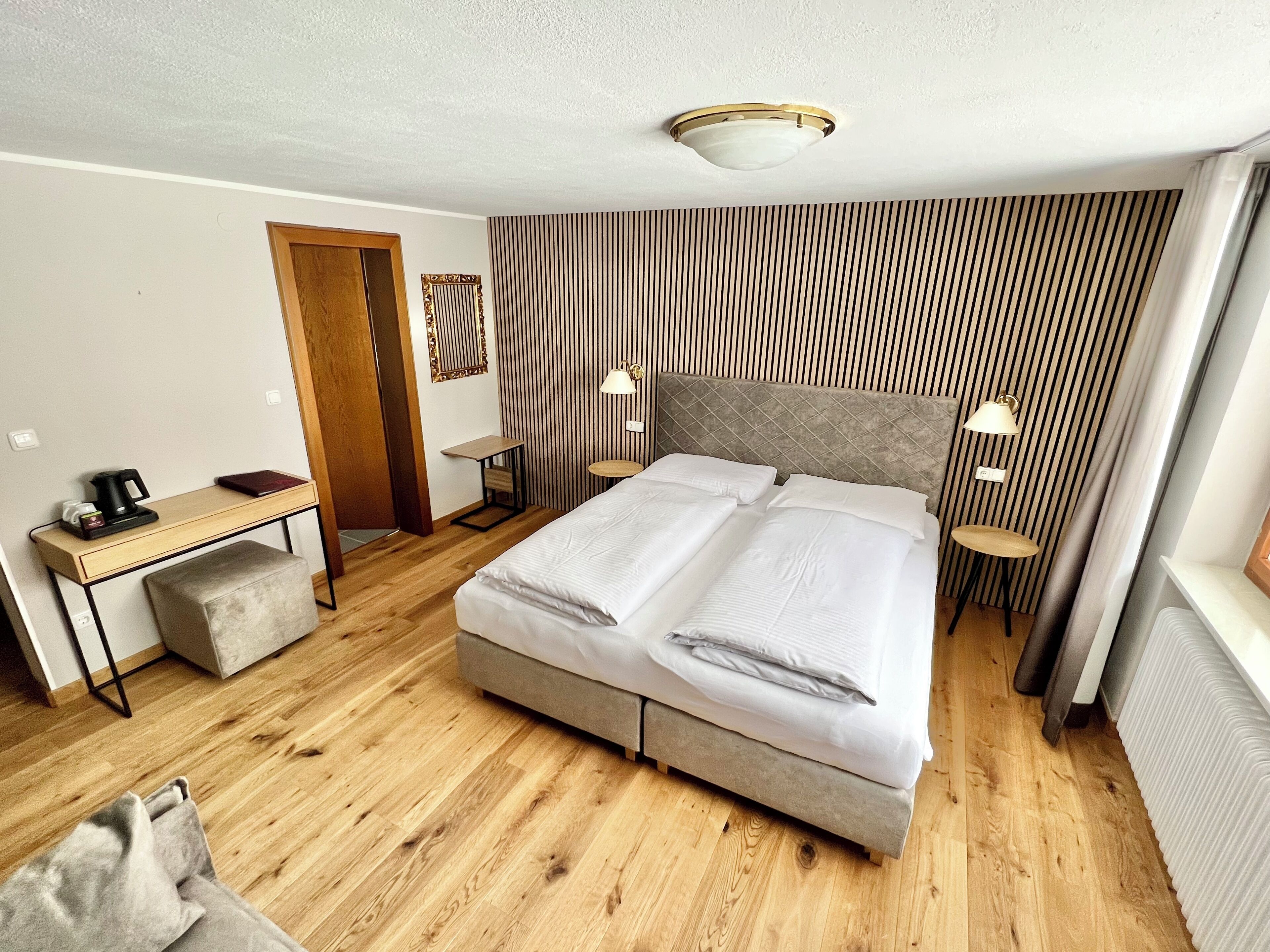 A hotel room with a bed, bedside lamps, a desk with a coffee maker, a small stool, and a striped wall.