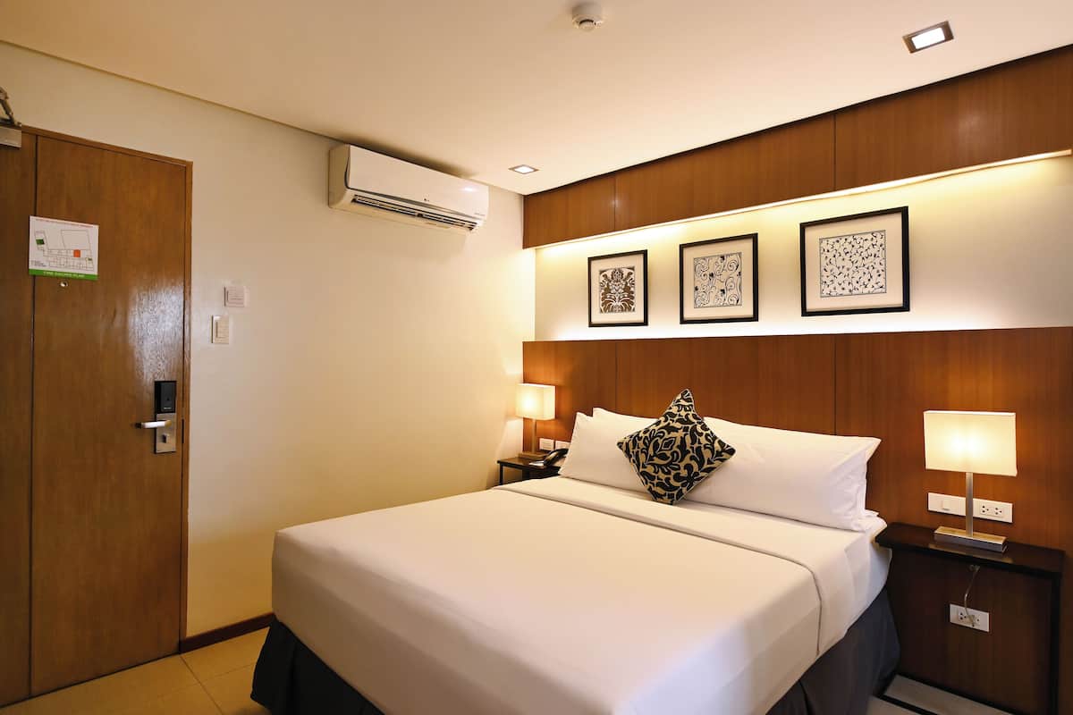 A hotel room with a large bed, wooden headboard, wall-mounted air conditioning, framed artwork, bedside lamps, and a door with a lock.