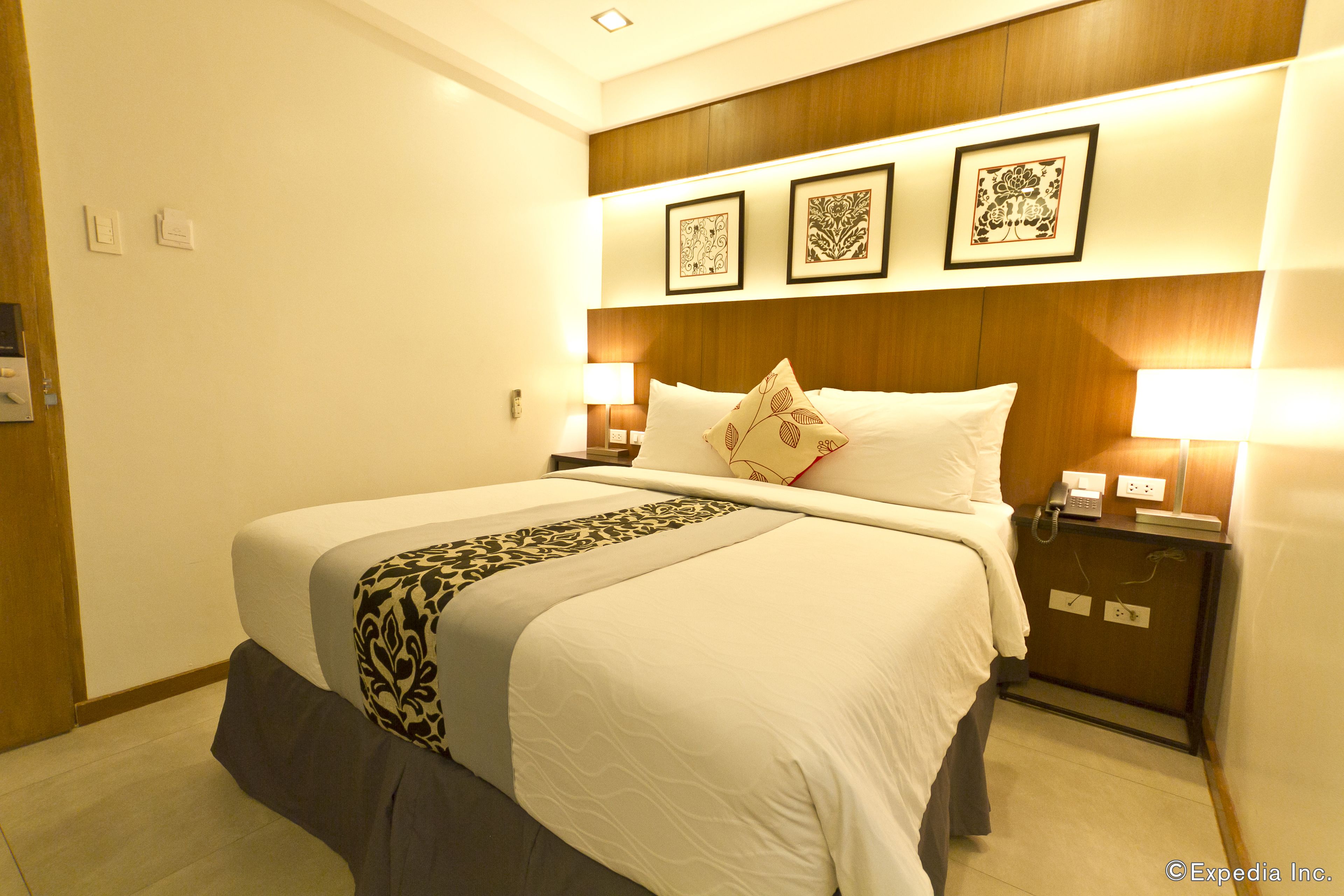 Una habitación de hotel con una cama grande, dos mesitas de noche con lámparas, un cabecero de madera y tres cuadros en la pared.