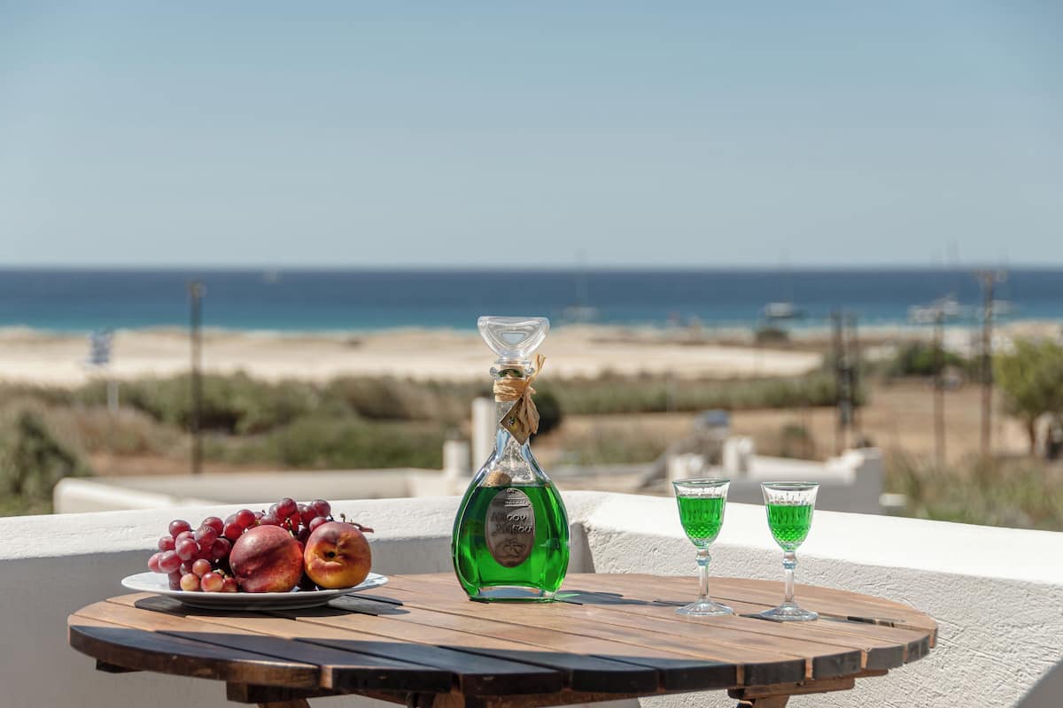 A table with a bottle of liquor, two glasses, and a plate of fruit by a beach view.