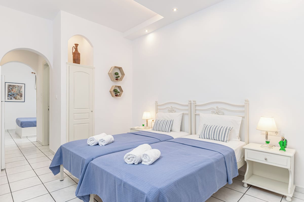 A hotel room with two beds, a white door, a small table with a lamp, and a wall-mounted shelf with decorative items.