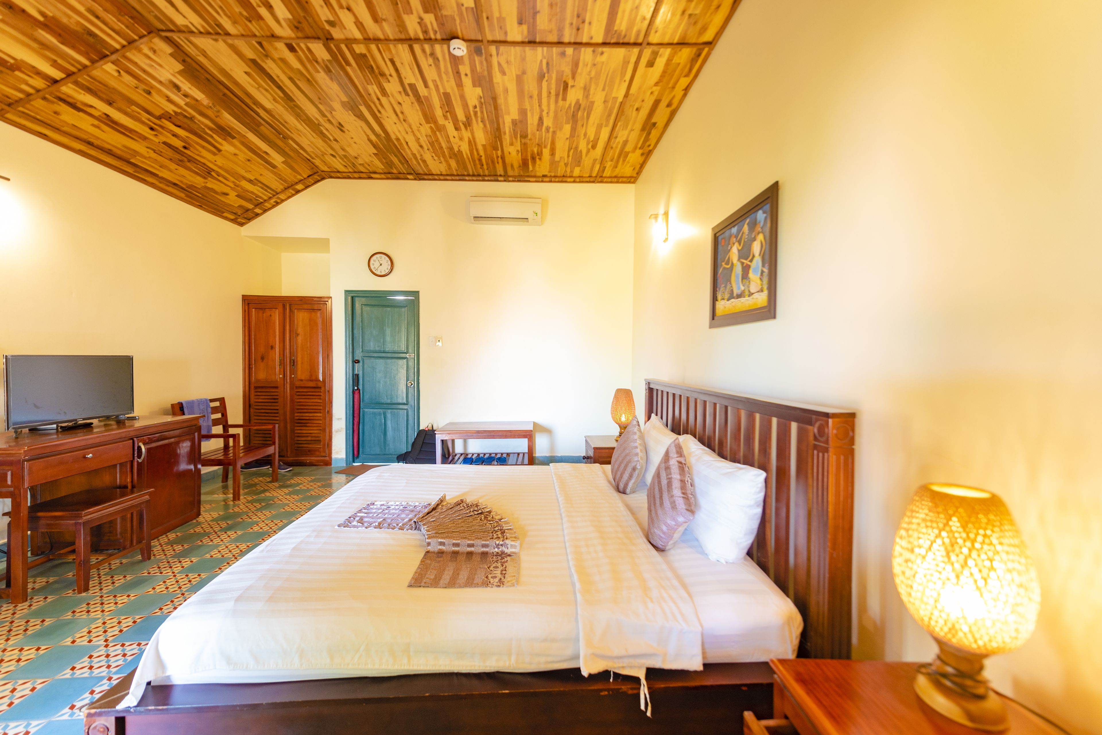 A hotel room with a wooden ceiling, a bed with white linens, a wooden desk with a television, a wooden wardrobe, and a wall-mounted air conditioner.