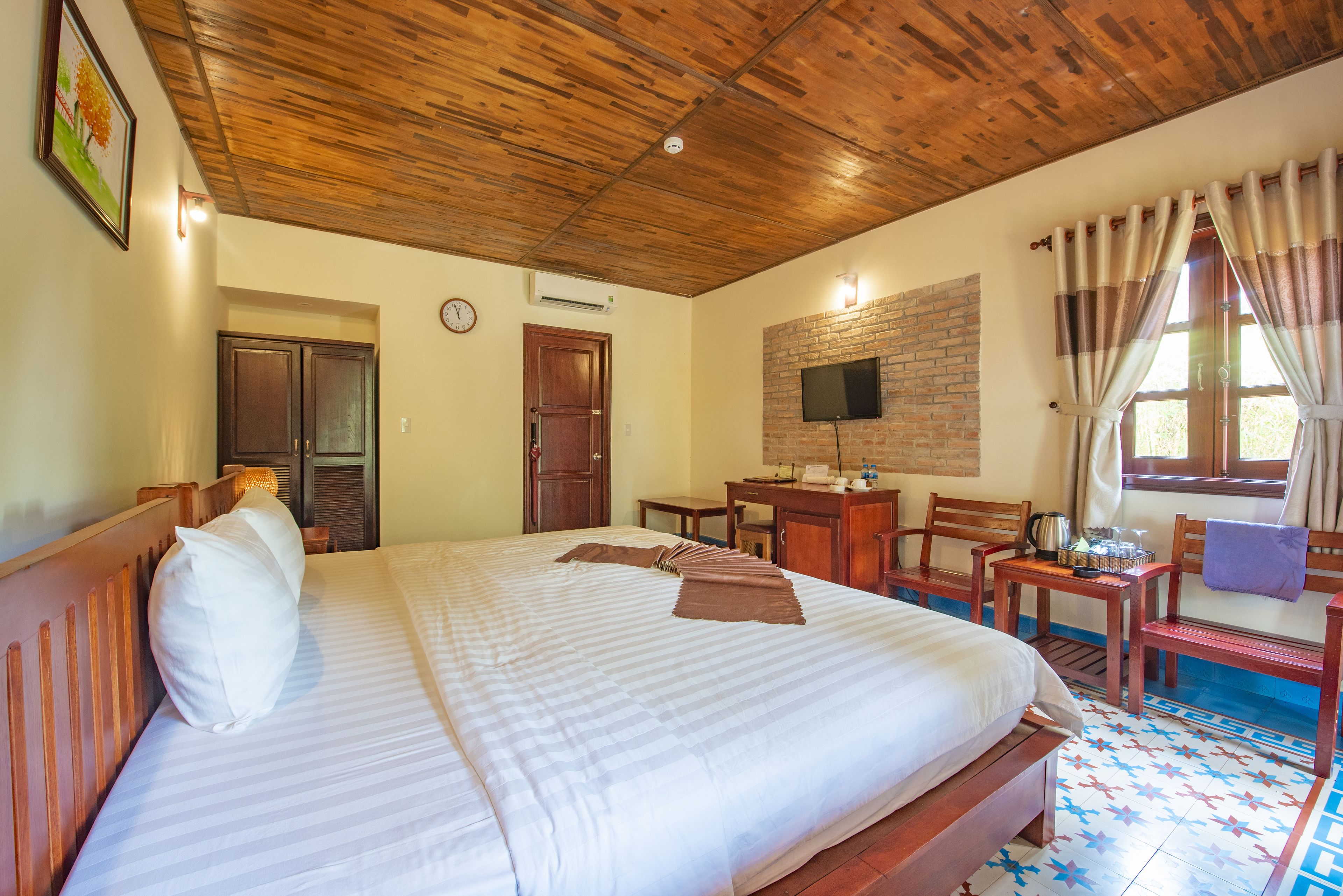 A hotel room with a large bed, wooden headboard, a TV mounted on a brick wall, a desk with a chair, and a window with curtains.