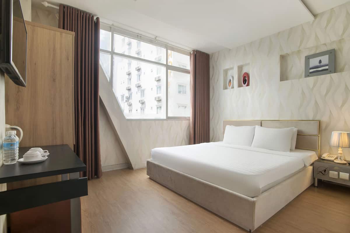 A hotel room with a large bed, a desk with a telephone, a water bottle, and a cup. There are curtains, a window with a view of buildings, and a wall with framed pictures.