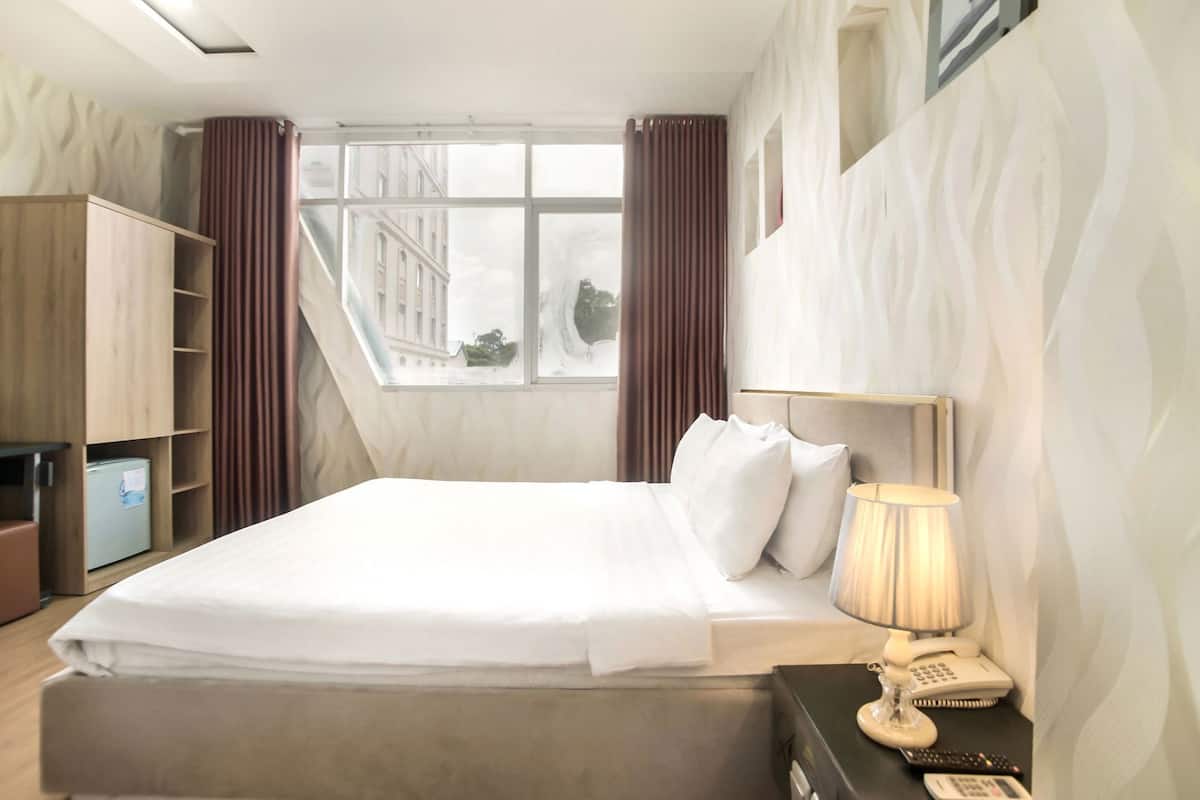 A hotel room with a bed, a desk with a lamp, a telephone, and a view of buildings through the window.