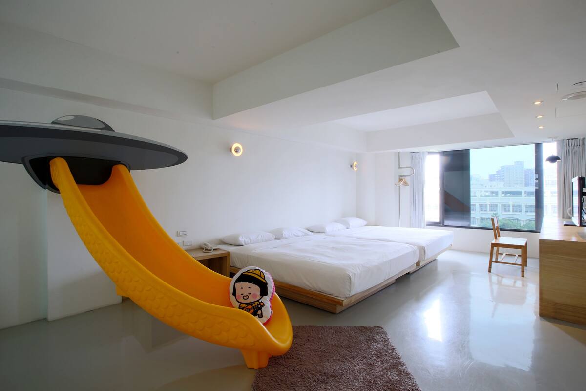 Un dormitorio moderno con cama, escritorio, silla, mesita pequeña, mesita de noche, ventana y un tobogán amarillo único.