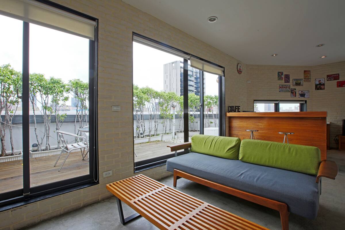 Una habitación moderna con sofá, banco y ventanas amplias que dan a un patio con árboles y un edificio.