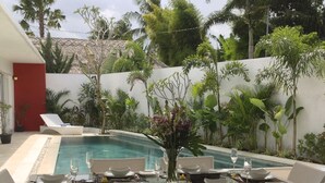 A poolside dining area with a table set for two, surrounded by wicker chairs, and a red door in the background. - Villa Sari Agung (Tibubeneng)
