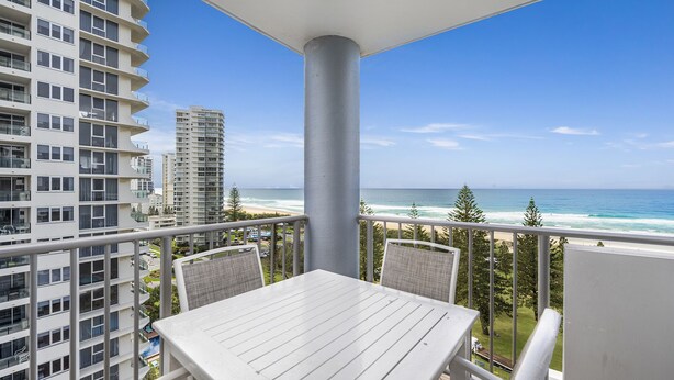 A balcony with a white table and chairs, overlooking a beach with ocean and tall buildings.