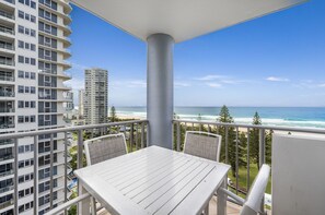 Un balcon avec une table et des chaises blanches, donnant sur une plage, l’océan et de grands immeubles.