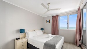 A bedroom with a bed, a nightstand, a ceiling fan, a window with curtains, and a view of the sea.