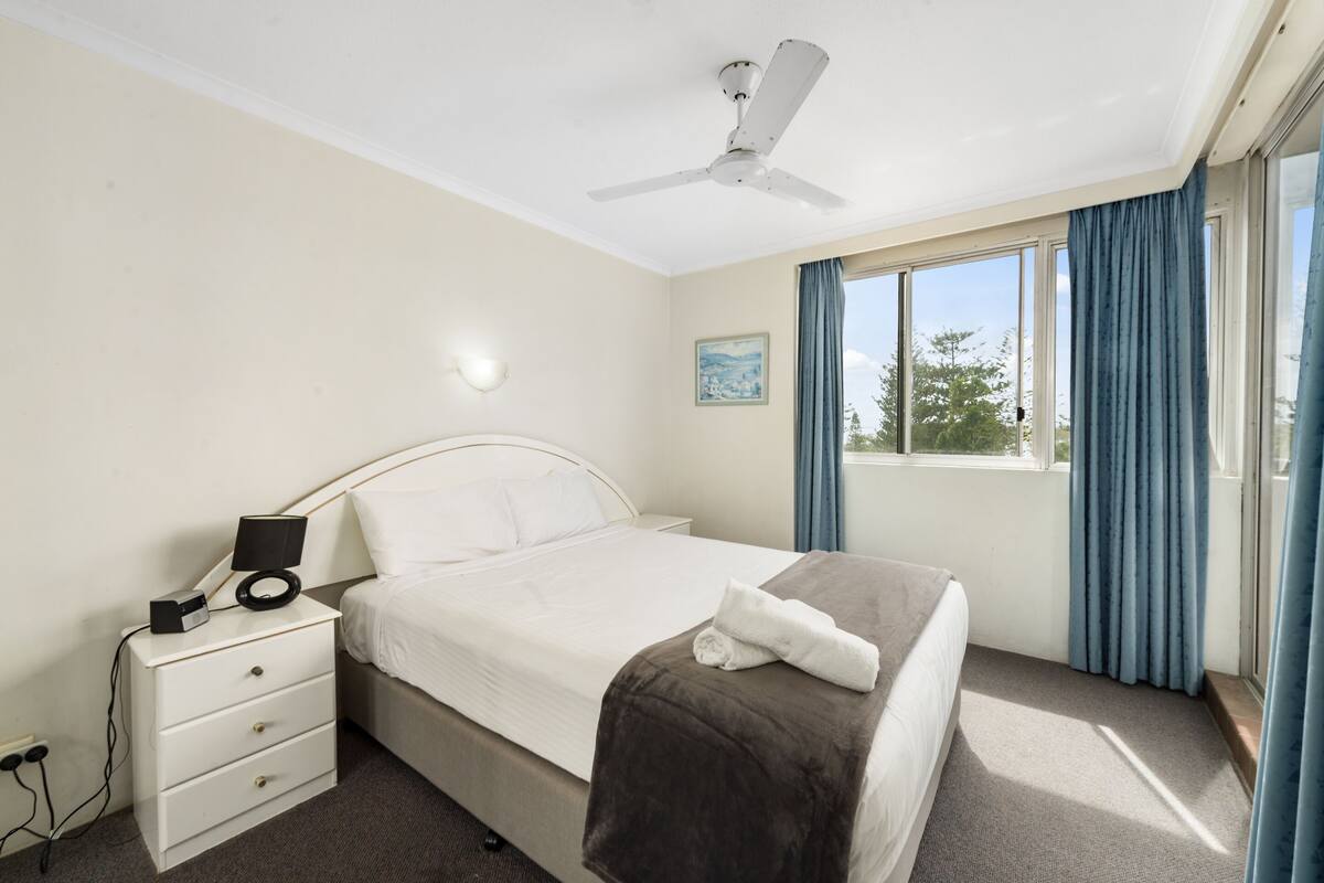 A hotel room with a bed, a bedside table with a lamp, a power outlet, and a window with curtains.