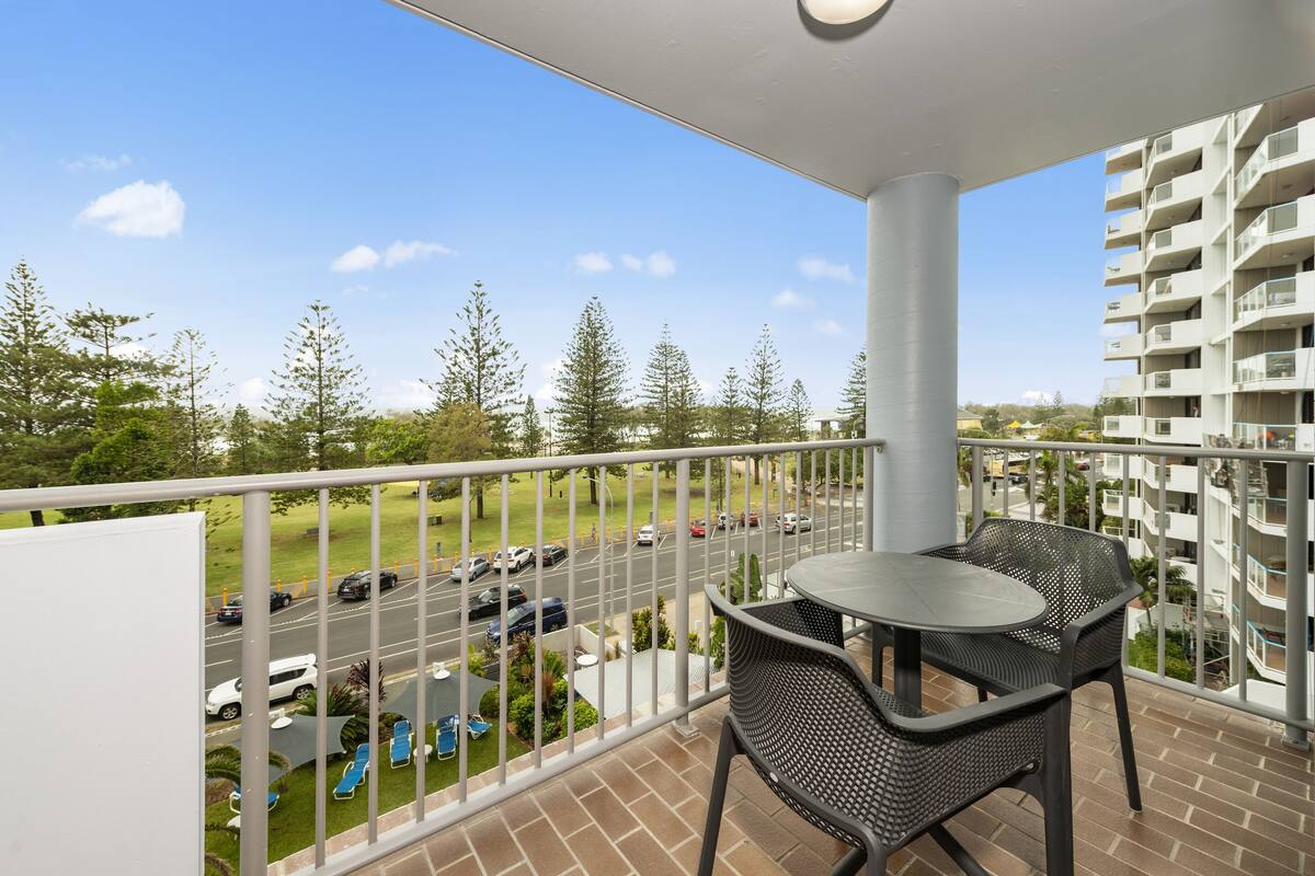 A balcony with a table and chairs, overlooking a street with cars and greenery.