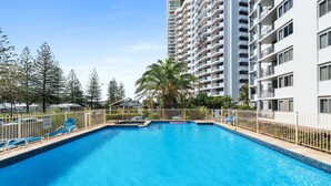 A swimming pool with lounge chairs, surrounded by a fence, and adjacent to a tall residential building.