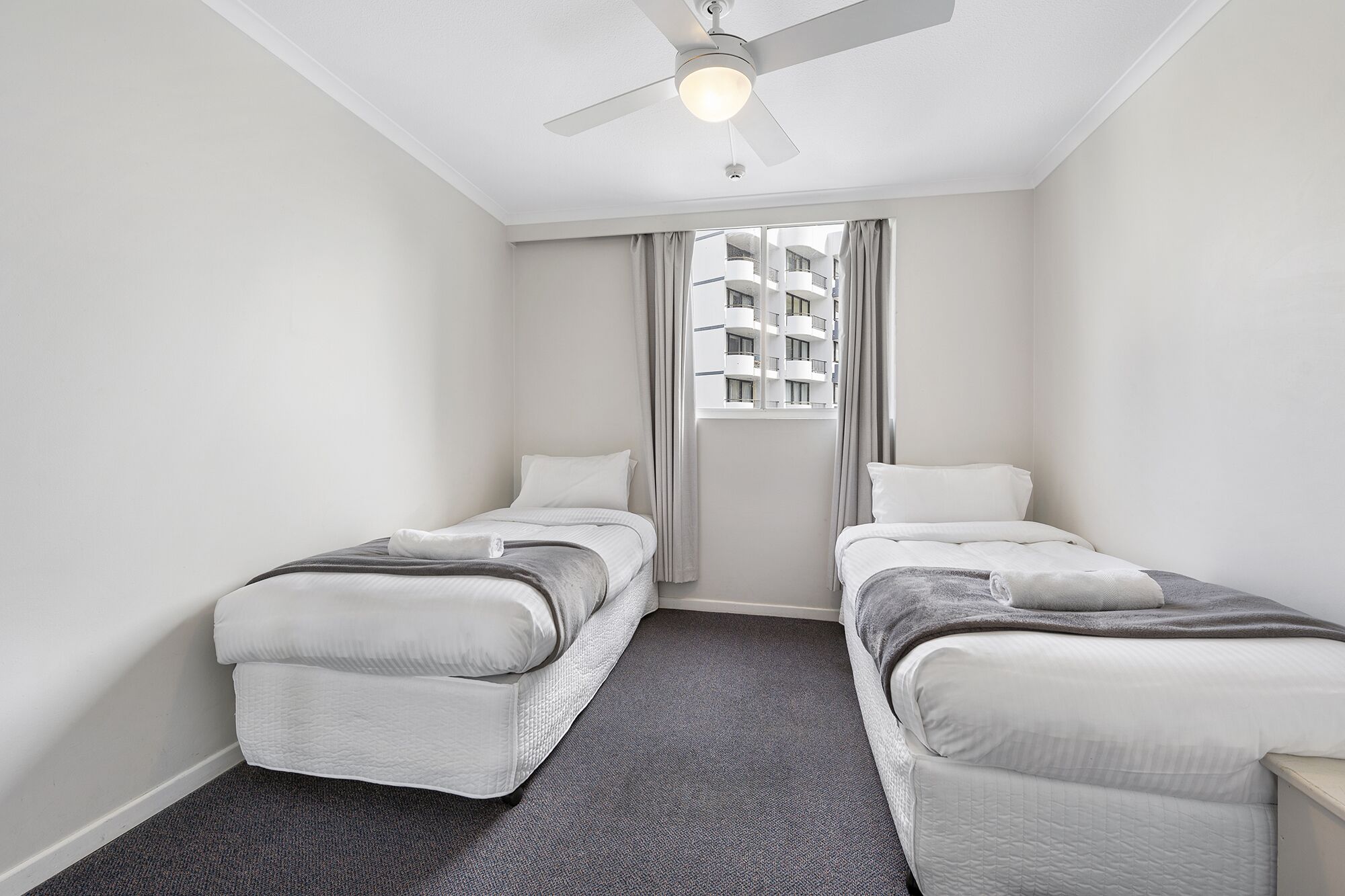 Ein Hotelzimmer mit zwei Einzelbetten, einem Deckenventilator, einem Fenster mit Vorhängen und Blick auf ein Gebäude.