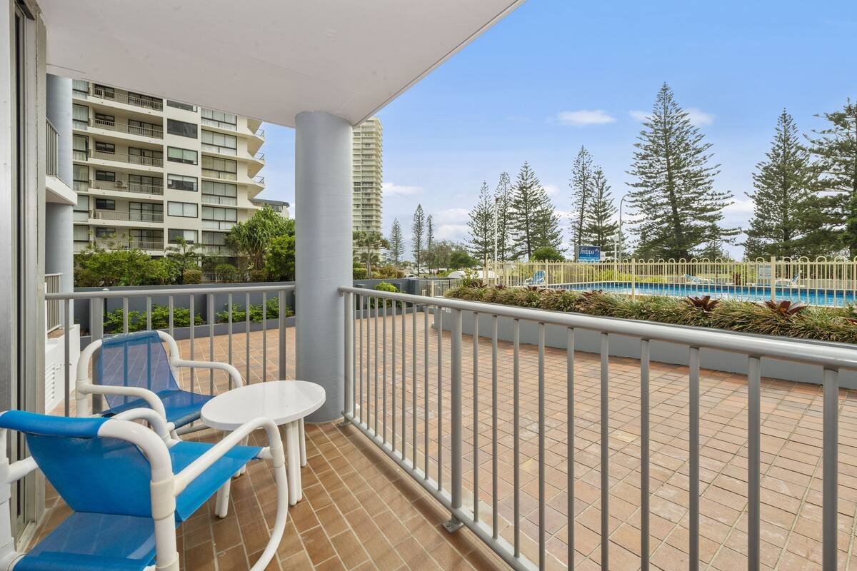 A balcony with two blue chairs and a small table, overlooking a swimming pool and tall buildings.