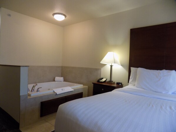 Habitación de hotel con una cama grande, un baño con bañera, lavamanos y una mesita de noche con lámpara.