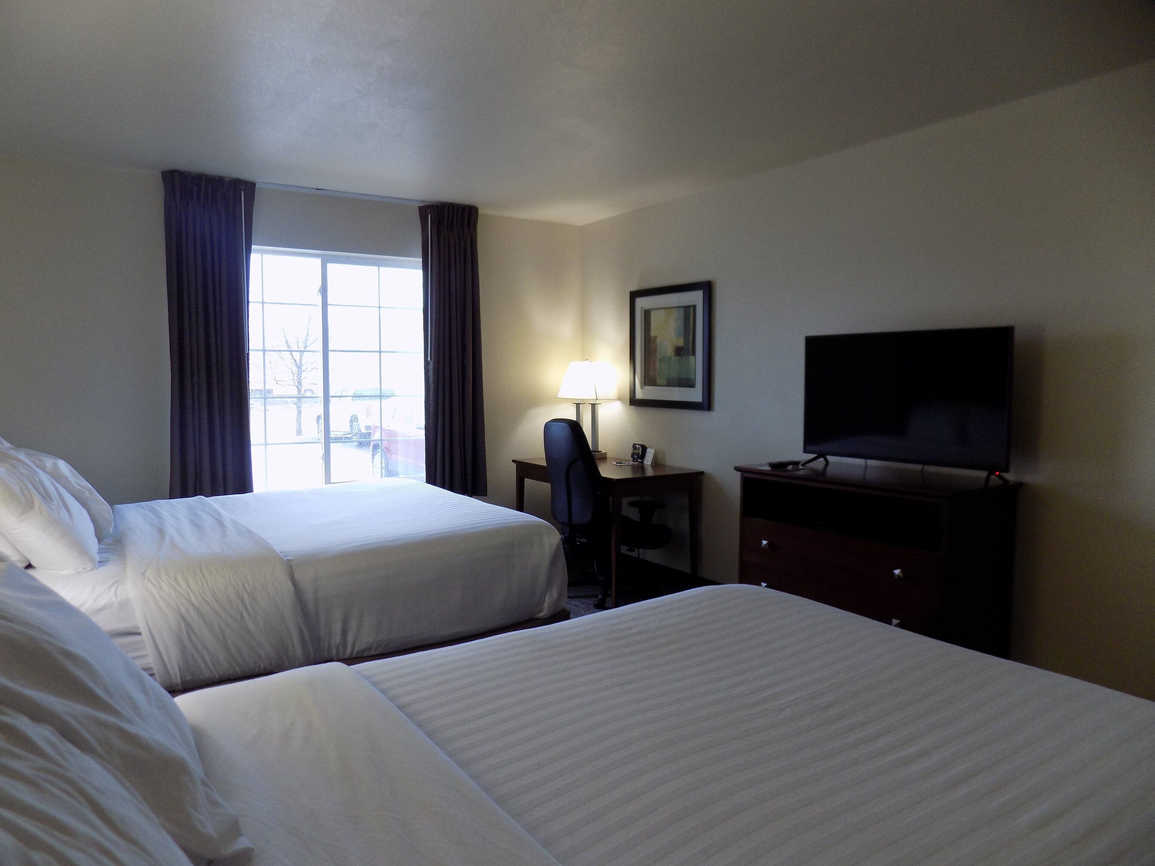 A hotel room with two beds, a desk with a chair, a TV, and a window with curtains.