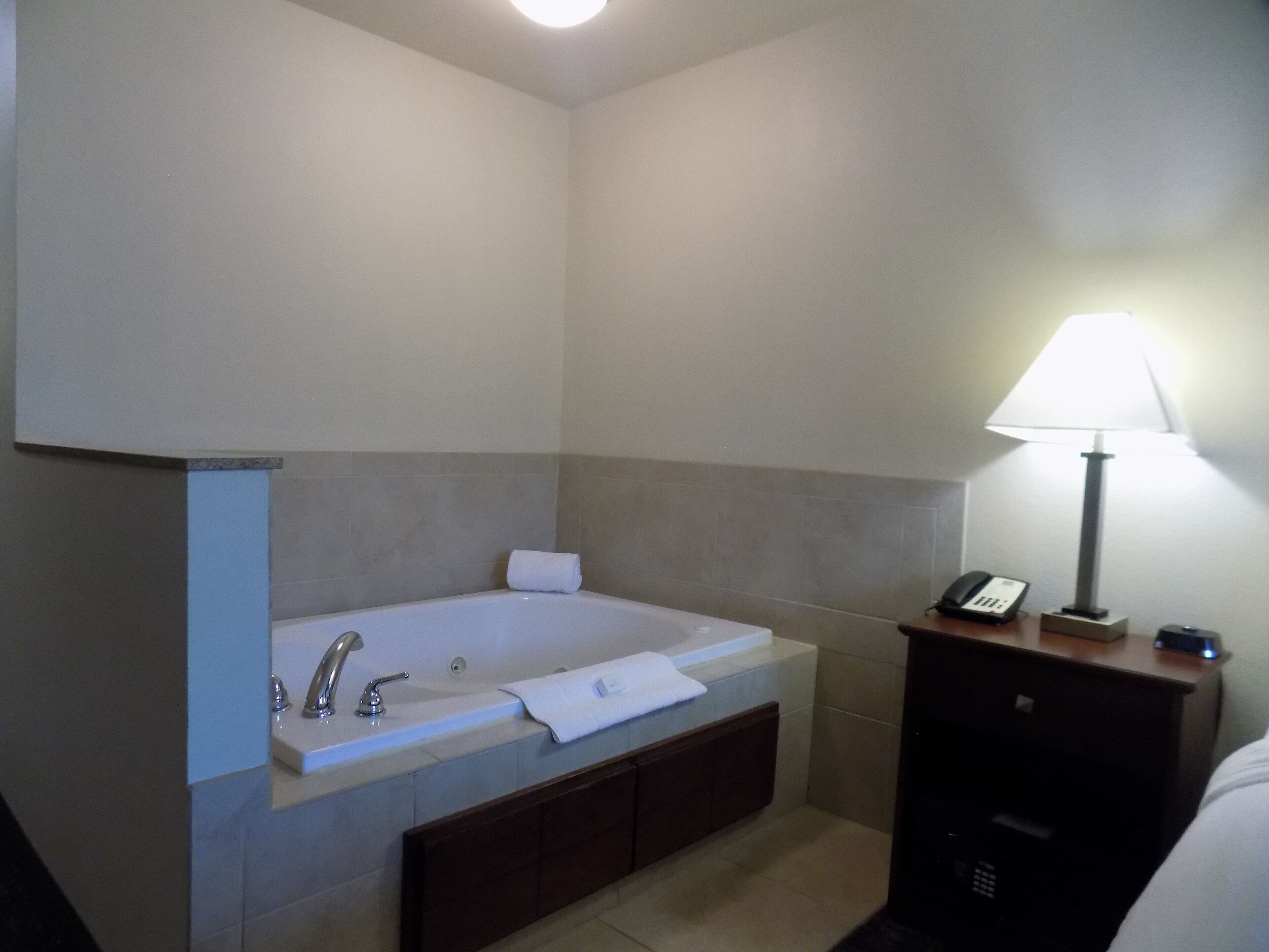A bathroom with a bathtub, a sink, a mirror, a towel, a nightstand with a lamp, and a phone.