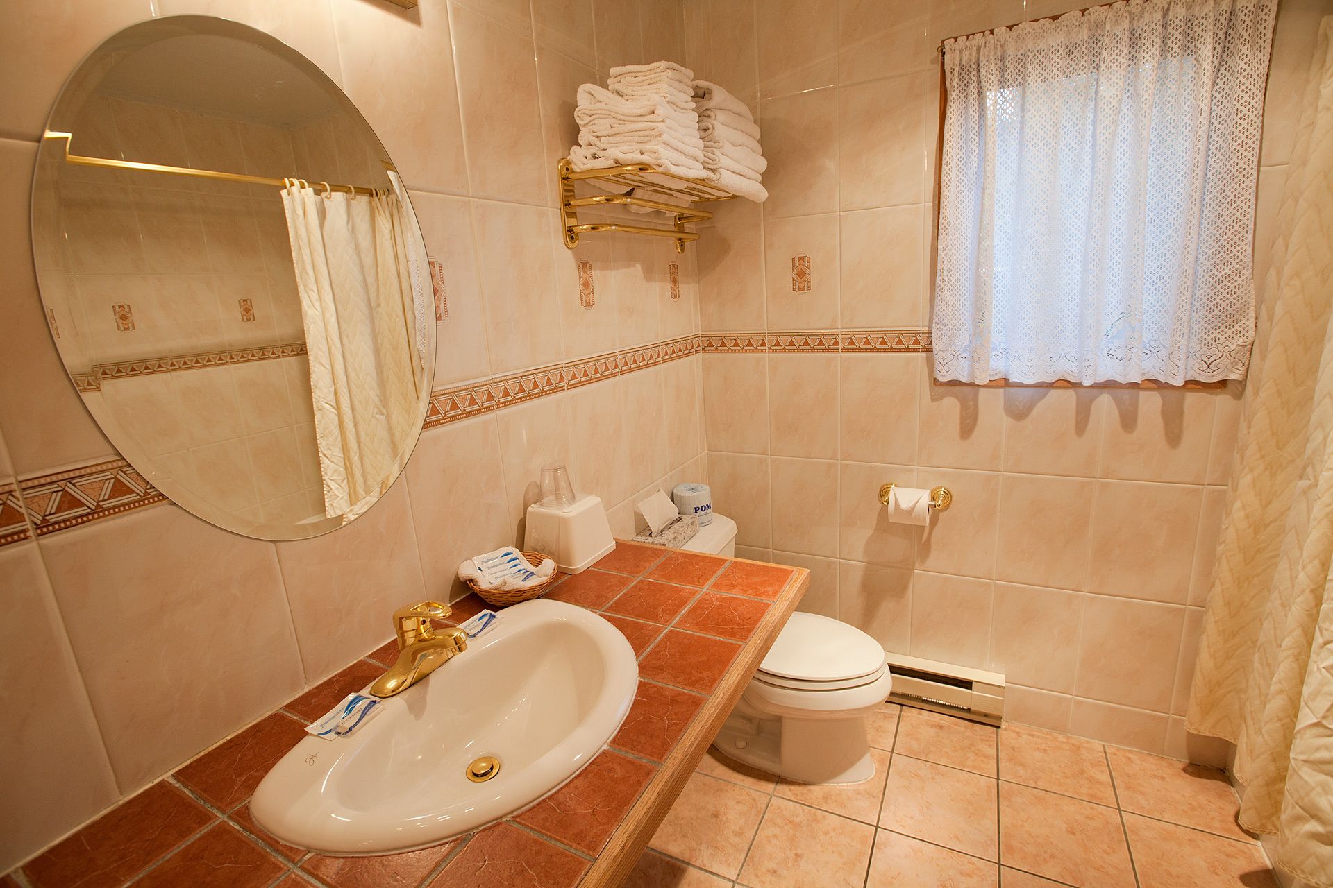 A bathroom with a sink, toilet, and a mirror. There are towels stacked on a shelf, a toilet paper holder, and a shower curtain.