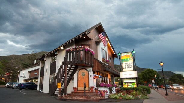 Um edifício de dois andares com fachada de madeira, varanda com flores e uma bandeira americana. Possui uma placa com a inscrição 'Starlight Lodge' e uma 'Loja de Presentes'.
