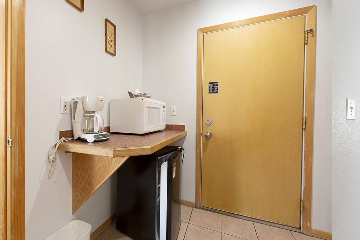 A hotel room with two beds, a small kitchenette, a wooden door, and a painting on the wall.