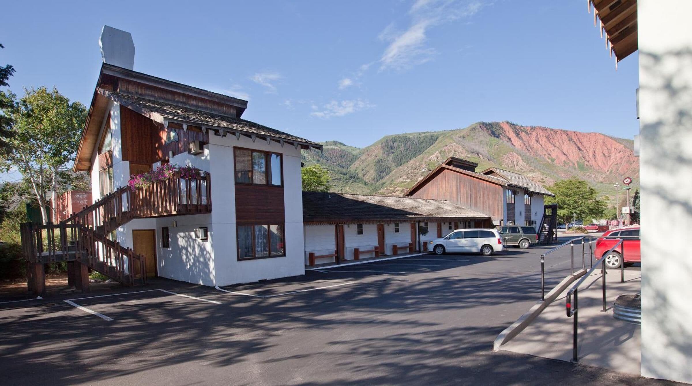 A building with a wooden facade and a balcony with flowers, a parking lot, and a mountain in the background.