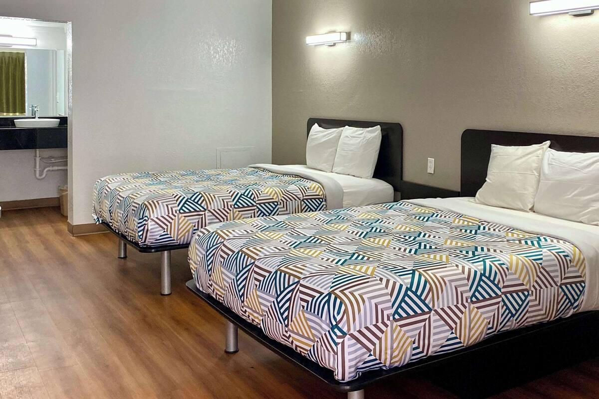 Zwei Einzelbetten mit gemusterten Bettbezügen, ein kleiner Nachttisch und ein Spiegel über dem Waschbecken in einem Hotelzimmer.