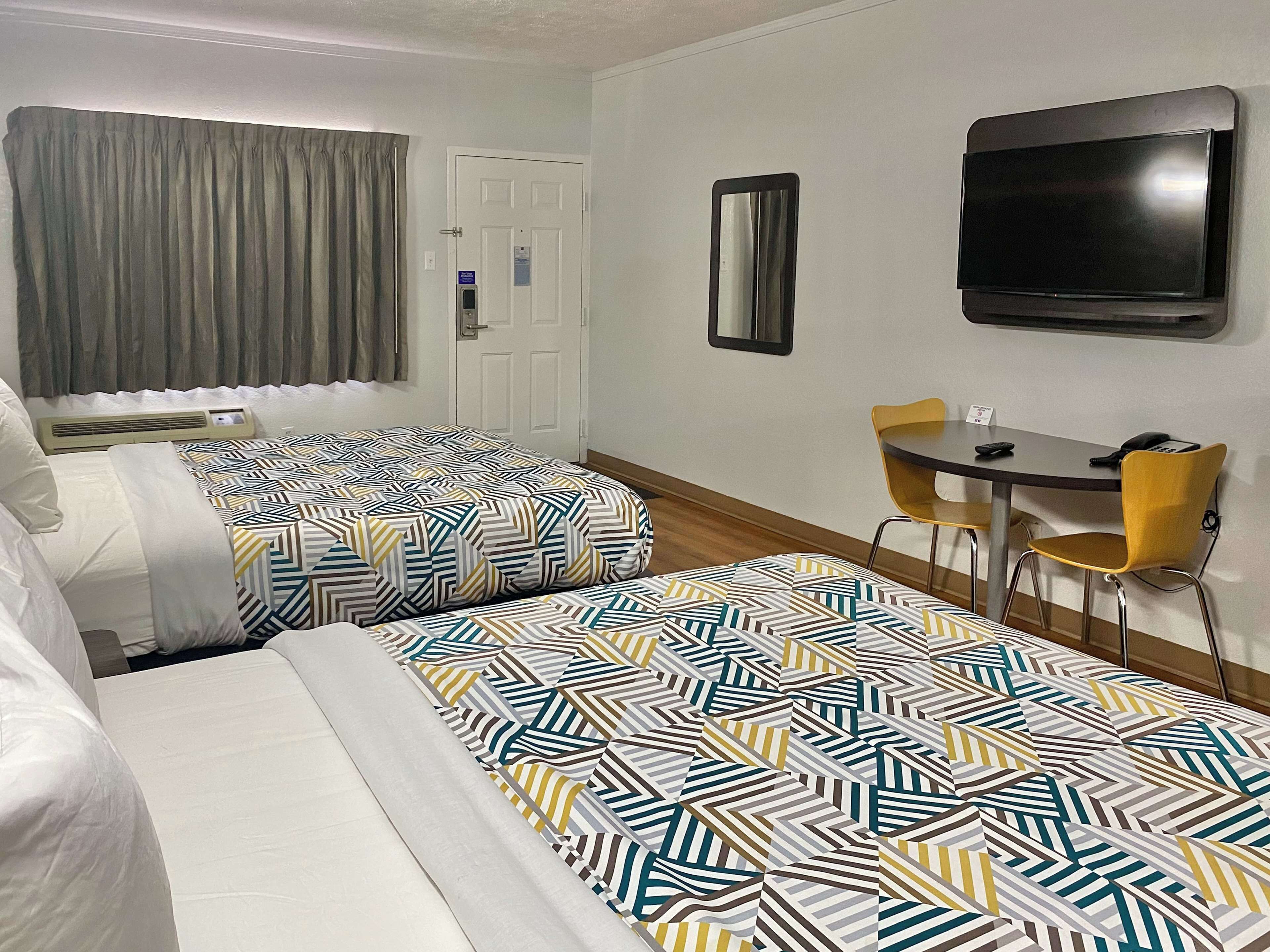 Quarto de hotel com duas camas, uma escrivaninha com cadeira, televisão e porta com placa.