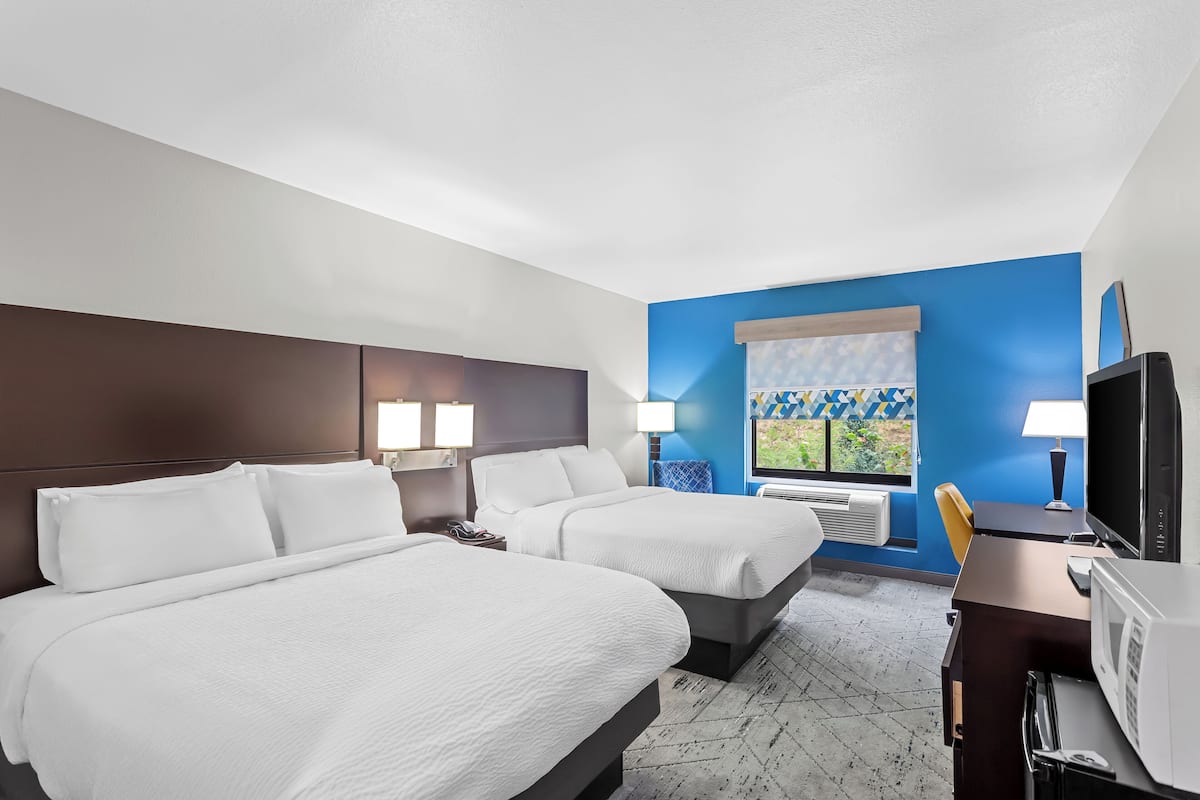 A hotel room with two beds, a desk, a TV, and a window with a view of greenery.
